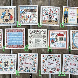 Könnte beinhalten: Eine Sammlung quadratischer dekorativer Fliesen mit Volkskunstmotiven. Jede Fliese zeigt farbenfrohe Illustrationen von Menschen, Häusern, Bäumen und Text. Die Fliesen sind auf einer Holzoberfläche angeordnet und präsentieren eine Vielzahl von Themen und Stilen.