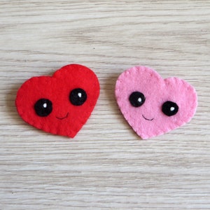 Broche en feutrine, coeur rouge ou rose avec de grands yeux noirs, et bouche souriante.