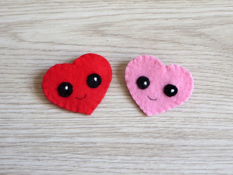 Broche en feutrine, coeur rouge ou rose avec de grands yeux noirs, et bouche souriante.