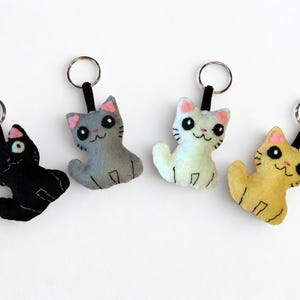 Op de afbeelding: Vier handgemaakte katvormige sleutelhangers in zwart, grijs, wit en geel. Elke pluche kat heeft roze oren, zwarte ogen en een lachend gezicht. De sleutelhangers hebben een zwarte lus en een zilveren metalen ring.