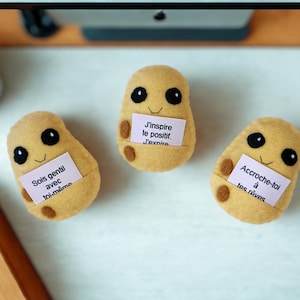 Peut inclure: Trois peluches en feutrine beige en forme de pomme de terre avec des taches marron foncé, des yeux brodés noirs et de petits sourires. Chaque peluche tient une petite carte rose pâle avec du texte : Sois gentil avec toi-même, J'inspire le positif.