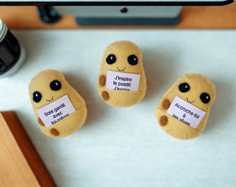 Peut inclure: Trois peluches en feutrine beige en forme de pomme de terre avec des taches marron foncé, des yeux brodés noirs et de petits sourires. Chaque peluche tient une petite carte rose pâle avec du texte : Sois gentil avec toi-même, J'inspire le positif.