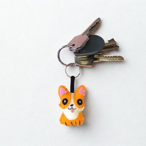 May include: A keyring featuring a small, felted corgi dog charm with orange and white fur, pink inner ears, and a black nose. The charm is attached to a silver metal split ring that holds a collection of various metal house and car keys.