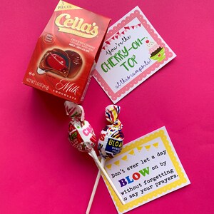 May include: A pink background with two lollipops, a box of chocolate cherries, and two cards with text. The text on the cards reads "You're the cherry-on-top at this campsite!" and "Don't ever let a day blow on by without forgetting to say your prayers."