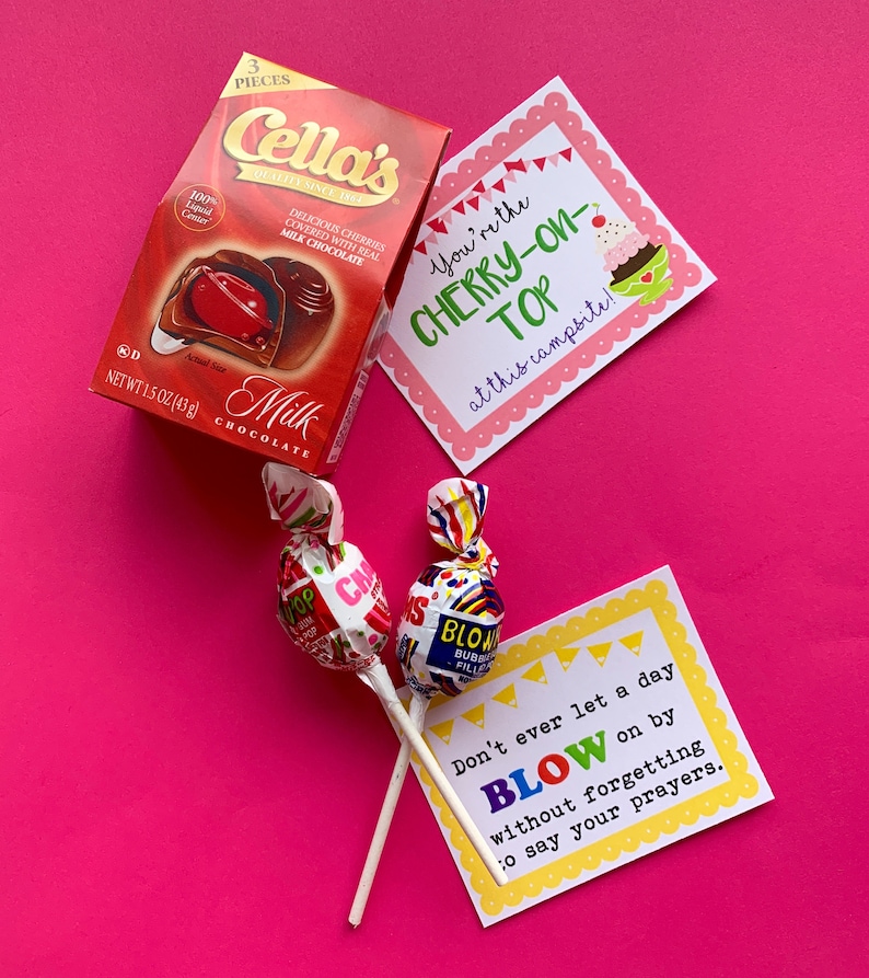 May include: A pink background with two lollipops, a box of chocolate cherries, and two cards with text. The text on the cards reads "You're the cherry-on-top at this campsite!" and "Don't ever let a day blow on by without forgetting to say your prayers."