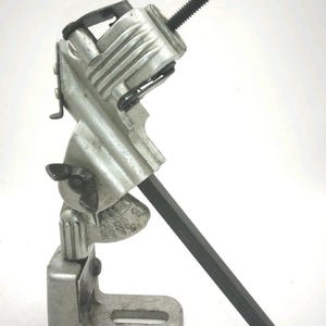 May include: A silver-colored metal adjustable mounting bracket featuring a base with a slotted hole, a central pivot point with a black wing nut, and an upper clamp mechanism with a threaded adjustment screw and a long black metal rod attached.