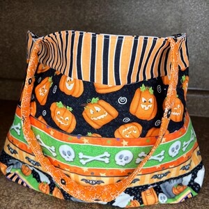 Könnte beinhalten: Eine Halloween-Tragetasche mit verschiedenen Mustern. Sie zeigt orange und schwarze Streifen, Kürbislaternen, Totenköpfe und Fledermäuse. Die Tasche hat orangefarbene Griffe und ist aus Stoff.