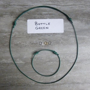 Pode incluir: Dois cordões verdes, um mais longo que o outro, com nós em cada extremidade. O cordão mais longo é um colar e o cordão mais curto é uma pulseira. Há três argolas de salto, uma prateada e duas douradas, no centro da imagem. Um cartão branco com o texto "Bottle Green" está na parte superior central da imagem.