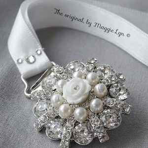 May include: A white satin ribbon sash featuring a silver-tone brooch with clear faceted crystals, round white pearls, and a central white floral accent. The text "The original, by Maggie Lyn ©" is printed on the ribbon in a black script font.