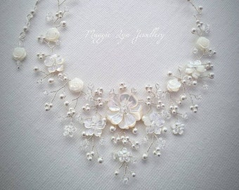 May include: A delicate white floral necklace with pearl and crystal accents. The necklace features a large, iridescent shell flower in the center.