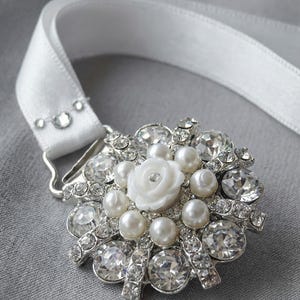 May include: A silver brooch with a white satin ribbon. The brooch features a central white rose surrounded by faux pearls and clear crystals. The ribbon is attached with small crystals. The brooch is a decorative item.
