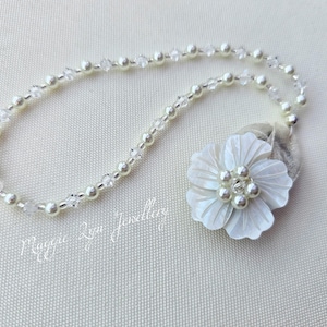 Puede incluir: Un collar de perlas blancas y cristales con un colgante de flor de concha blanca. La flor tiene un grupo de perlas blancas en el centro.