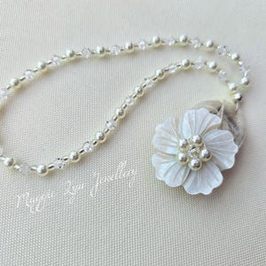 May include: A delicate necklace featuring a strand of small, round, white pearls and clear crystals. A large, white, flower-shaped pendant with pearl accents and a fabric backing is attached. The text "Maggie Lyn Jewellers" is visible.