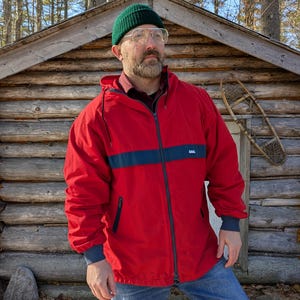 May include: A red hooded jacket with a dark blue stripe across the chest and dark blue cuffs. The jacket is zipped up and worn with blue jeans and a green beanie. The background features a log cabin and trees.