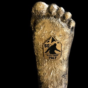 May include: A gold-colored, textured, foot-shaped sculpture with a gold plaque that reads "50th Bluff Creek 1967". The plaque features a silhouette of a person standing on a mountain.