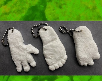 May include: Three white, textured, keychains shaped like a hand, a foot, and a foot. Each keychain has a silver chain attached.