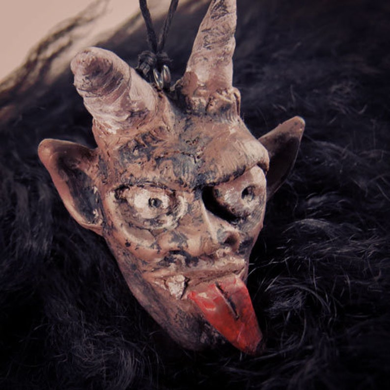 Peut inclure: Un pendentif en c&eacute;ramique brune en forme de t&ecirc;te de diable avec des cornes, une langue pointue et une surface textur&eacute;e. Le pendentif est suspendu &agrave; un cordon noir.