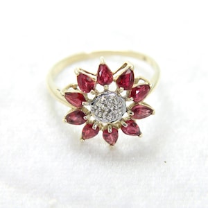 Puede incluir: Un anillo de oro con un diseño floral. Los pétalos están formados por piedras preciosas de color rojo intenso, que rodean un centro de pequeñas piedras blancas brillantes. El anillo está sobre un fondo blanco.
