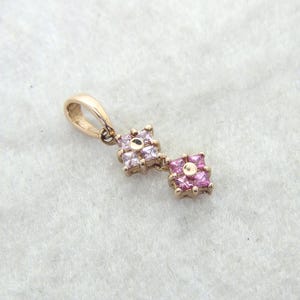 A gold pendant with two square floral clusters. Each cluster is made of small, pink gemstones. The pendant has a loop for a chain. The background is a light, textured surface.