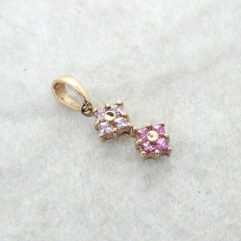 A gold pendant with two square floral clusters. Each cluster is made of small, pink gemstones. The pendant has a loop for a chain. The background is a light, textured surface.