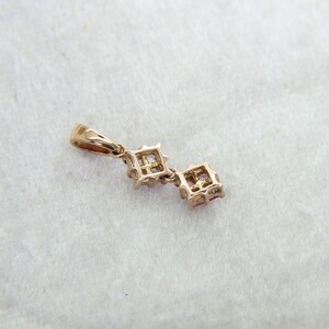 May include: A gold-toned pendant featuring two square-cut pink stones set in a metal frame. The pendant includes a small bail at the top for attachment to a chain, showcasing a delicate design with visible metal prongs holding each stone.