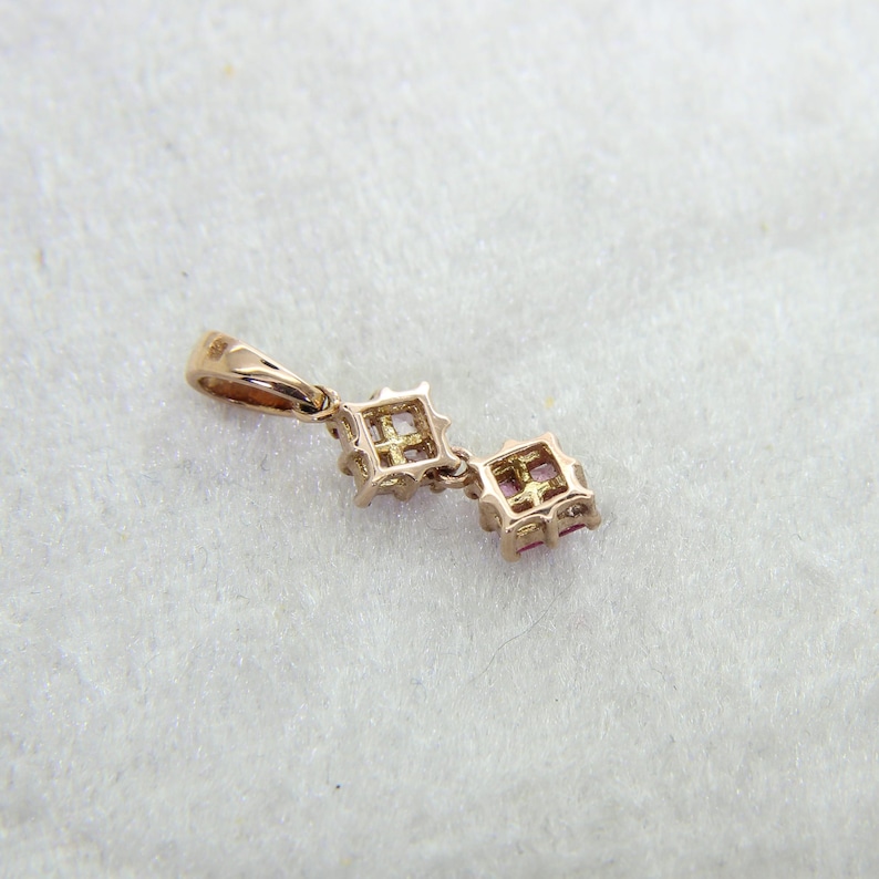 May include: A gold-toned pendant featuring two square-cut pink stones set in a metal frame. The pendant includes a small bail at the top for attachment to a chain, showcasing a delicate design with visible metal prongs holding each stone.