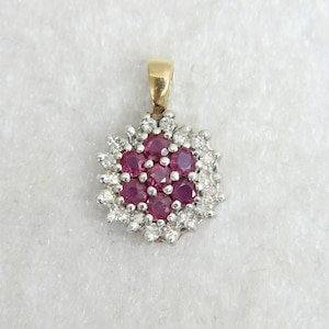 Puede incluir: Un colgante de oro con un grupo de piedras preciosas redondas de color rojo intenso, rodeadas de piedras preciosas más pequeñas y transparentes. El colgante tiene una argolla de oro para sujetarlo a una cadena. Las piedras están engastadas en un metal plateado.