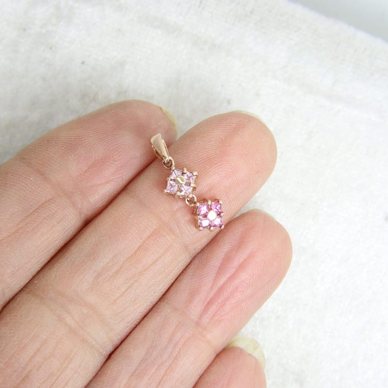 May include: A rose gold pendant featuring two square-shaped links, each set with four small pink stones arranged in a cluster around a central point. The pendant includes a small metal bail at the top for attachment to a chain or necklace.