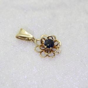 May include: A gold pendant featuring a flower design with a dark blue gemstone center. The pendant has a delicate, openwork petal design and a small loop for a chain. The metal has a polished finish.