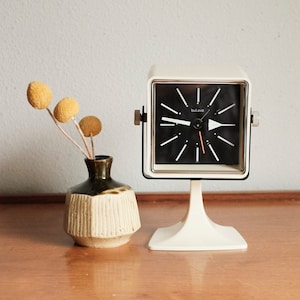 Op de afbeelding: Een vintage witte Bulova wekker met een zwarte wijzerplaat en zilveren accenten. De klok staat op een witte standaard. Links van de klok staat een kleine vaas met gedroogde bloemen. De klok heeft een retro-ontwerp.