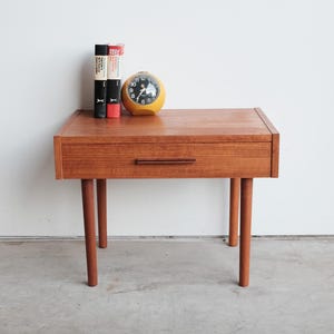 May include: A mid-century modern wooden nightstand with a single drawer and four tapered legs. A yellow alarm clock and a stack of books sit on top. The nightstand is made of wood and has a warm, natural finish.