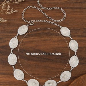Pu&ograve; includere: Cintura argentata con elementi decorativi ovali. La cintura ha una chiusura a catena ed &egrave; lunga circa 70 cm, con altri 48 cm di catena. Presentata su una superficie di legno.