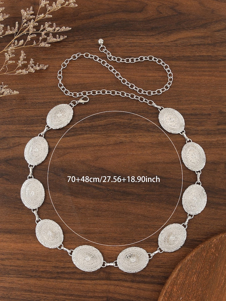 Pu&ograve; includere: Cintura argentata con elementi decorativi ovali. La cintura ha una chiusura a catena ed &egrave; lunga circa 70 cm, con altri 48 cm di catena. Presentata su una superficie di legno.