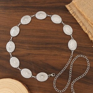 Pu&ograve; includere: Cintura color argento con medaglioni ovali con intricati disegni in rilievo. La cintura &egrave; collegata da piccoli anelli e include un'estensione a catena per la regolazione della taglia. Esposta su una superficie di legno.