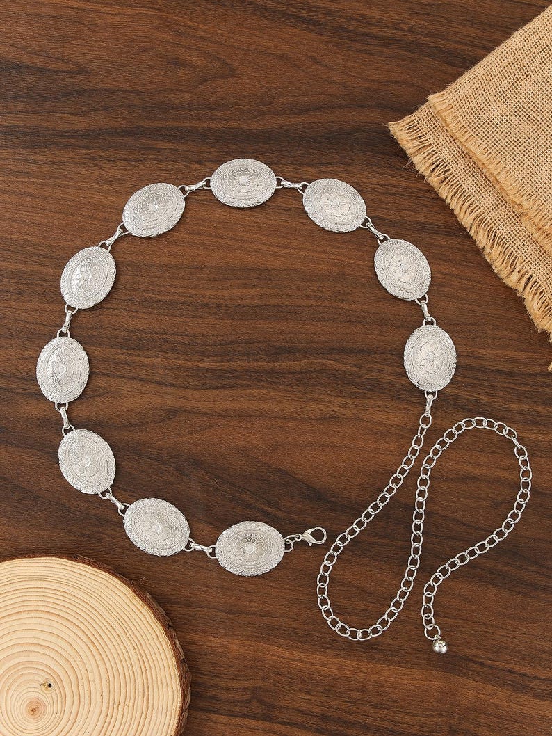 Pu&ograve; includere: Cintura color argento con medaglioni ovali con intricati disegni in rilievo. La cintura &egrave; collegata da piccoli anelli e include un'estensione a catena per la regolazione della taglia. Esposta su una superficie di legno.