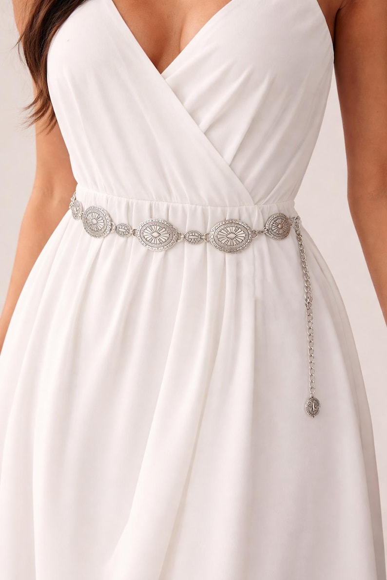 May include: Silver-coloured metal belt with oval and circular decorative elements. The belt features an intricate design and a chain with a decorative pendant. The belt is worn over a white dress.