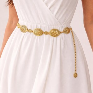 May include: Gold-coloured metal belt with oval and circular decorative elements. The belt features an intricate design and a dangling chain with a matching charm. The belt is worn over a white dress, adding a touch of elegance to the outfit.