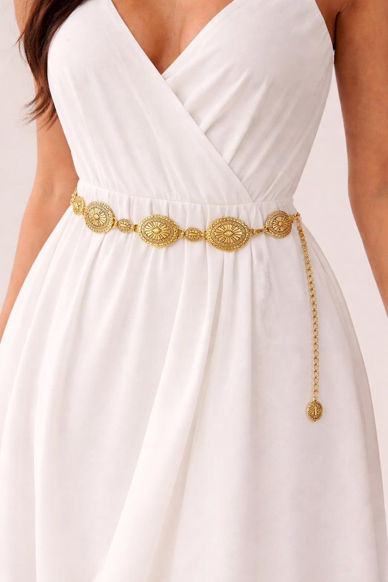 May include: Gold-coloured metal belt with oval and circular decorative elements. The belt features an intricate design and a dangling chain with a matching charm. The belt is worn over a white dress, adding a touch of elegance to the outfit.