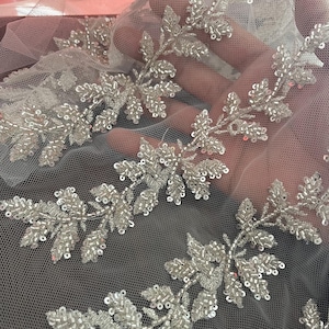 May include: Close-up of a delicate, silver beaded floral lace trim. The trim features intricate leaf and flower designs, with each element meticulously crafted with small, shiny beads. The lace is set against a sheer, light-coloured fabric, creating a shimmering effect.