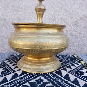 May include: A gold metal container with an ornate design and a lid. The container has a round base and a tall, pointed top. The container is sitting on a patterned fabric.