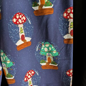 May include: Dark blue fabric with a repeating pattern of mushroom-shaped snow globes. Each globe features a colorful mushroom house with a red or green cap, a wooden base, and a dusting of white snow. The design is whimsical and festive.