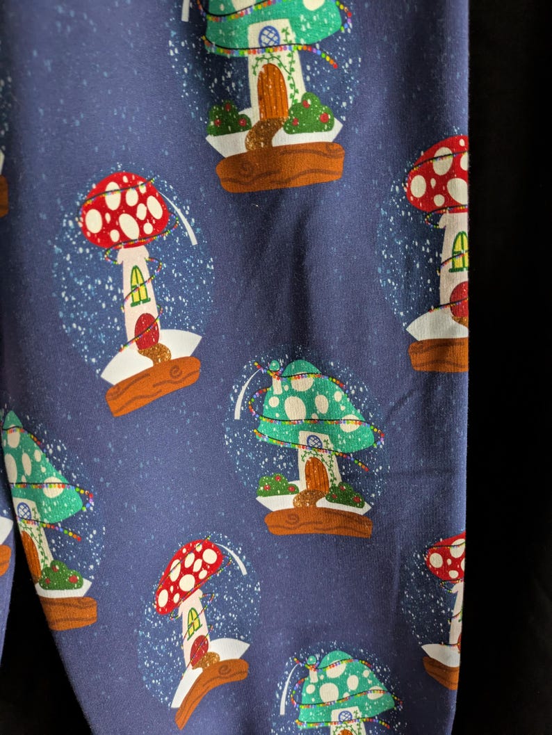 May include: Dark blue fabric with a repeating pattern of mushroom-shaped snow globes. Each globe features a colorful mushroom house with a red or green cap, a wooden base, and a dusting of white snow. The design is whimsical and festive.