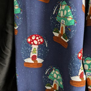 May include: Dark blue fabric with a repeating pattern of mushroom-shaped snow globes. The snow globes have red and green mushroom caps with white polka dots, and are set on brown bases. White flecks simulate falling snow.