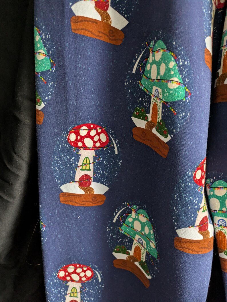 May include: Dark blue fabric with a repeating pattern of mushroom-shaped snow globes. The snow globes have red and green mushroom caps with white polka dots, and are set on brown bases. White flecks simulate falling snow.