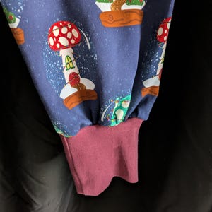 May include: Close-up of a blue fabric with a repeating pattern of whimsical mushroom houses. The design features red and white mushroom caps, green foliage, and brown bases. The cuff is a solid burgundy color.