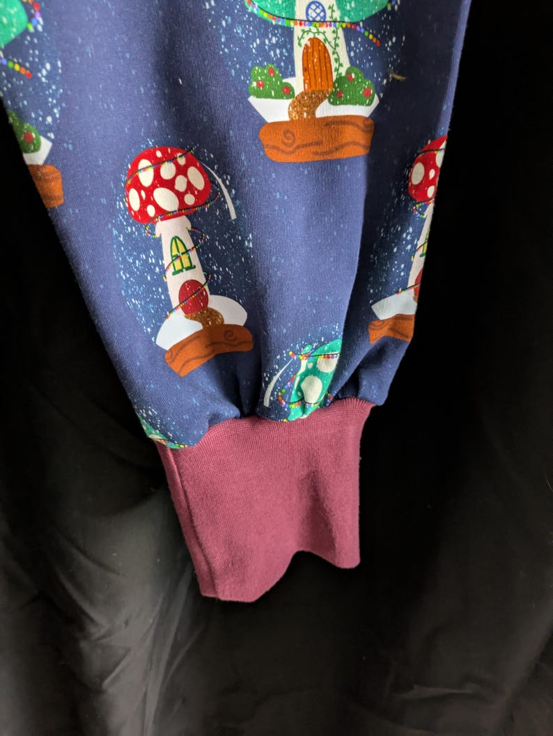 May include: Close-up of a blue fabric with a repeating pattern of whimsical mushroom houses. The design features red and white mushroom caps, green foliage, and brown bases. The cuff is a solid burgundy color.