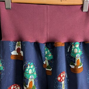 May include: Close-up of a pair of pants with a dark blue background and a pattern of colorful mushroom houses. The waistband is a solid mauve color. The text "Bobtail's Boutique" is visible on the wooden hanger.