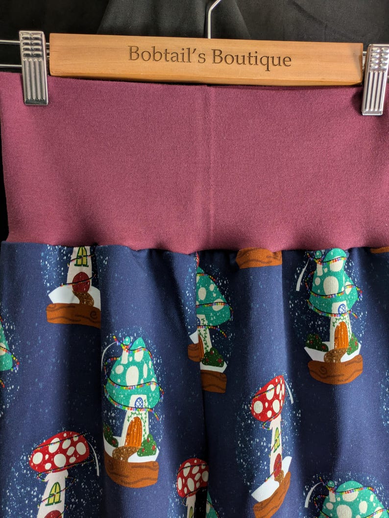May include: Close-up of a pair of pants with a dark blue background and a pattern of colorful mushroom houses. The waistband is a solid mauve color. The text "Bobtail's Boutique" is visible on the wooden hanger.