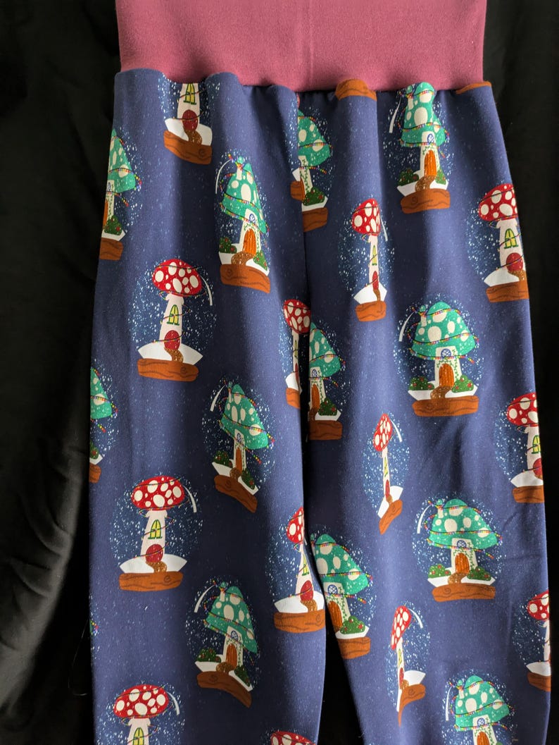 May include: Blue pants with a repeating pattern of mushroom-shaped snow globes. The mushrooms have red and green caps, with small houses inside the globes. The waistband is a solid mauve color.