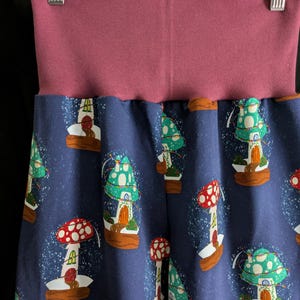 May include: Blue pants with a repeating pattern of mushroom houses in red, green, and white. The waistband is a solid mauve color. The pants are hanging on a wooden hanger with the text "Bobtail's Boutique".